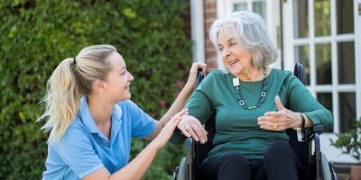 Carer Pushing Senior Woman In Wheelchair Outside Home